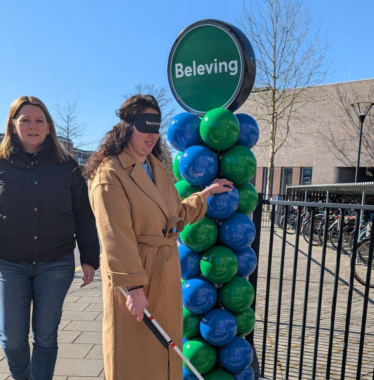 Vrouw loopt naast andere vrouw langs ingang van de beleving, met taststok en oogmasker.