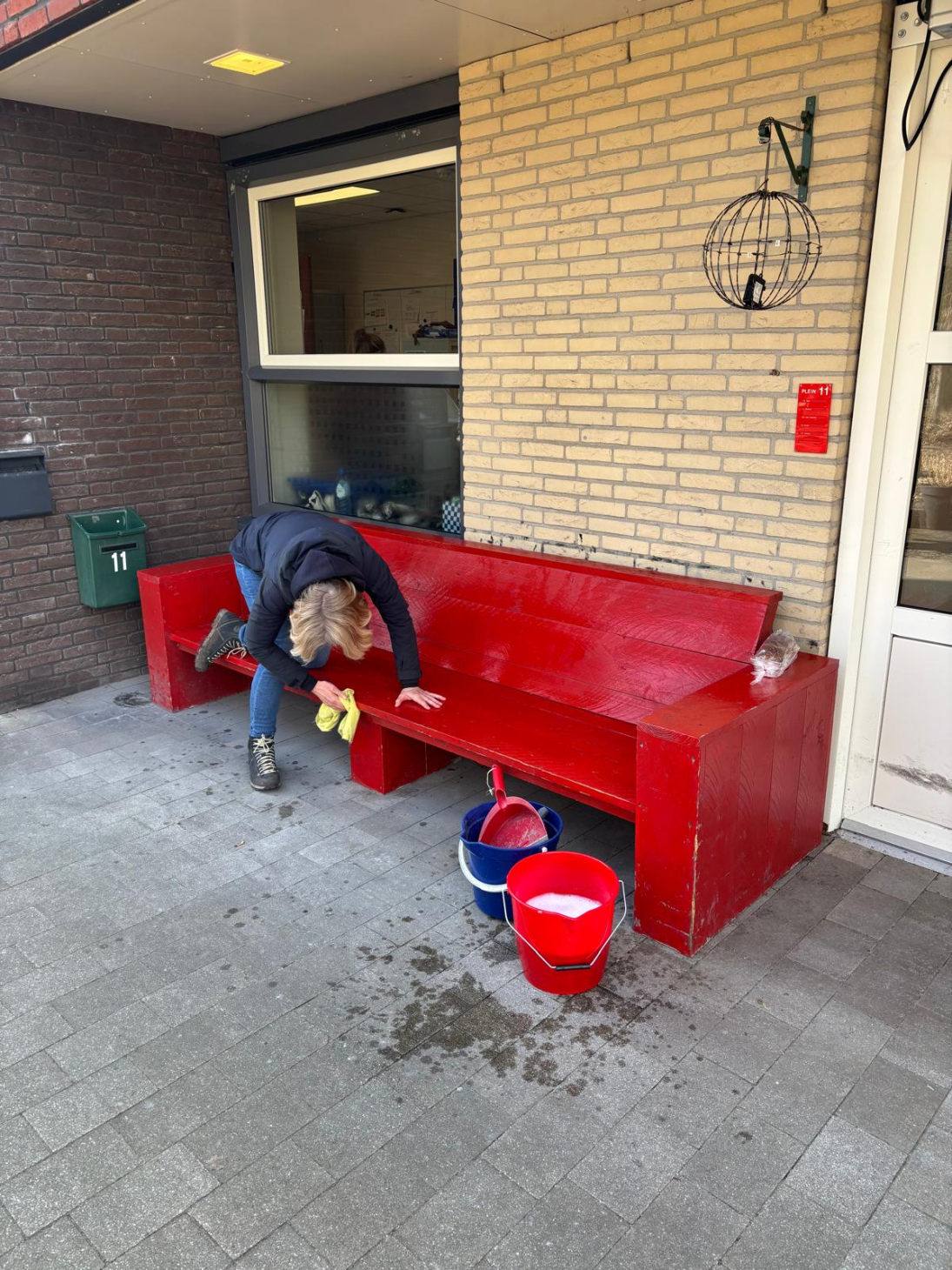 Carola maakt een rode, houten bank schoon die voor een woning staat