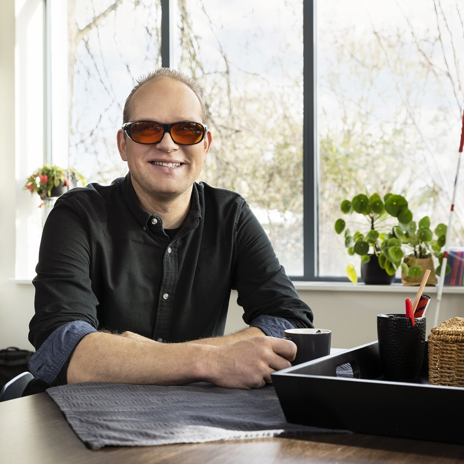 Afbeelding van een man aan een bureau. Met zonnebril en kopje koffie in zijn handen. Hij kijkt in de camera.