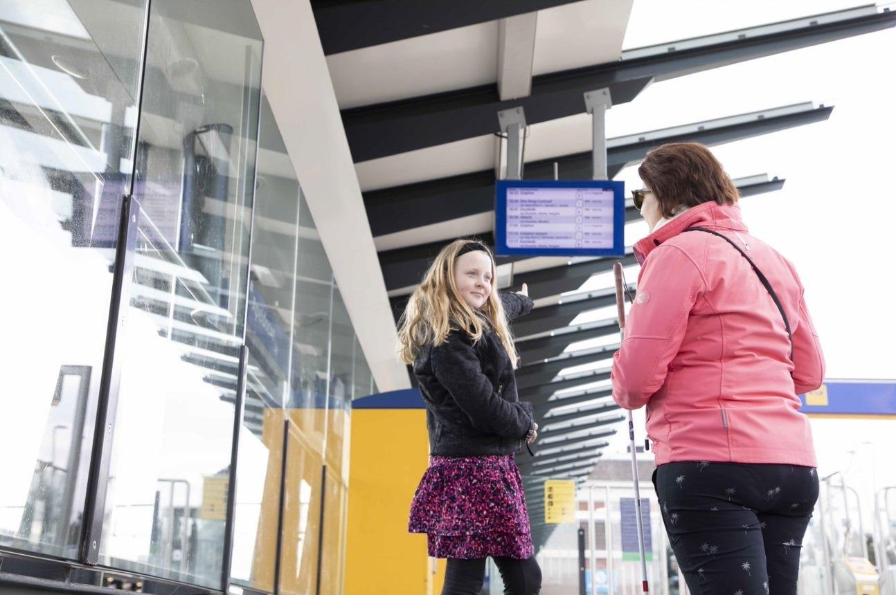 Jong blond meisje staat met haar moeder voor een stationsbord