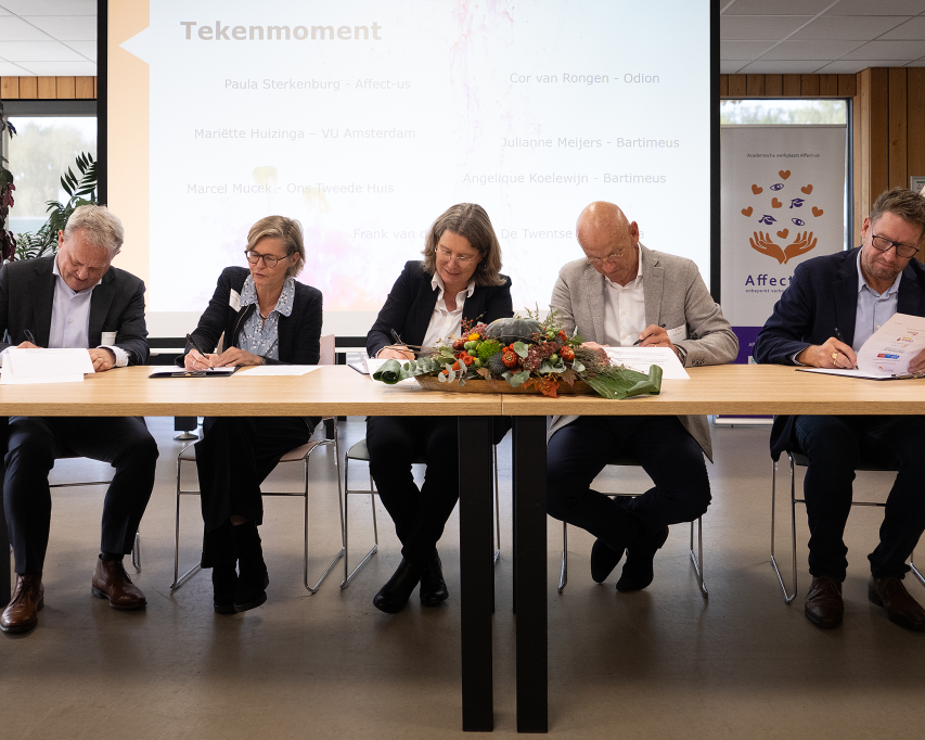 5 mensen op rij aan tafel die papieren ondertekenen