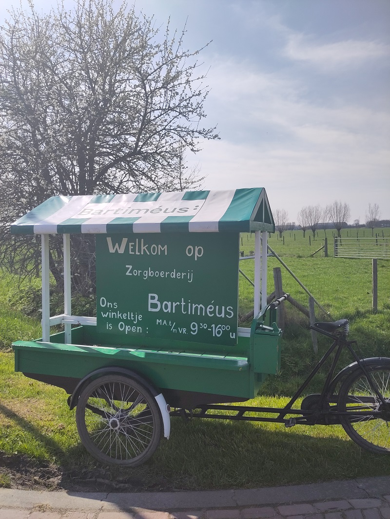 Een fietskar bij de ingang van Boerderij Langbroek. Op een groen bord staat: Welkom op de Zorgboerderij Bartiméus. Het winkeltje is open van maandag t/m vrijdag van 09.00 tot 16.00 uur.