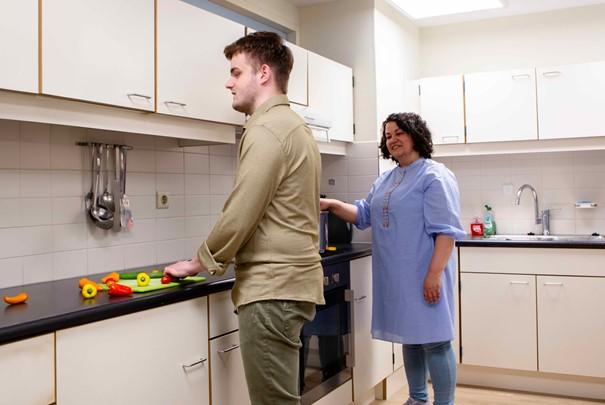 Een jongeman snijdt groenen in de keuken en naast hem staat een begeleider die toekijkt