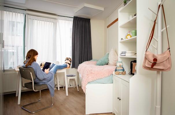 Een dame zit in haar kamer op een stoel met haar voeten omhoog op het bureau en ze leest een boek