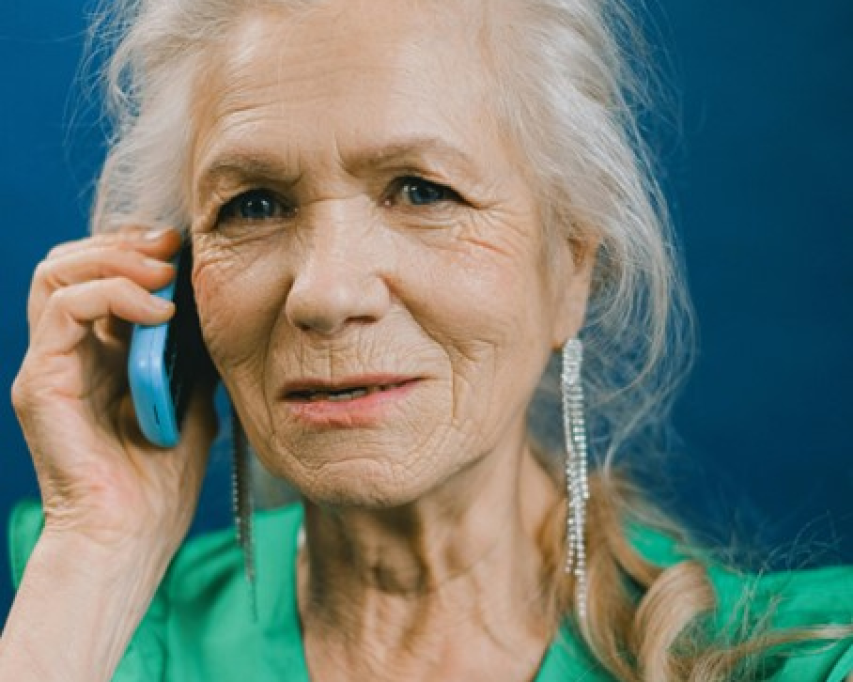Een dame met grijs haar en groene blouse houdt een telefoon met blauw hoesje tegen haar oor en kijkt in de camera